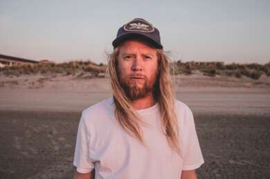 Stu Larsen steht am Strand. Er trägt ein weißes T-Shirt und eine graue Mütze.