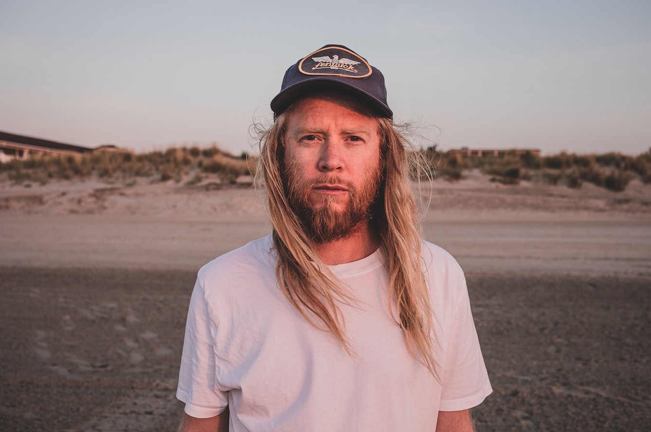 Stu Larsen steht am Strand. Er trägt ein weißes T-Shirt und eine graue Mütze.