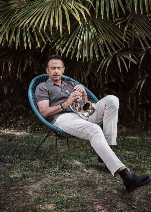 Till Brönner sitzt mit seiner Trompete in den Händen auf einem Stuhl. Im Hintergrund sind Palmen zu sehen.
