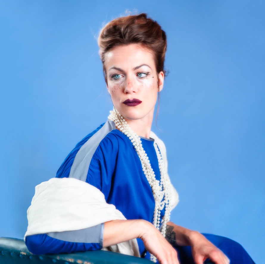 Charlotte Brandi sitzt in einem blauen Kleid vor einem hellblauen Hintergrund