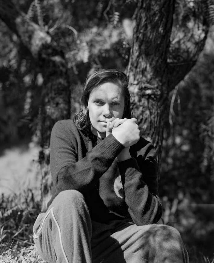 Rhye sitzt in einer Schwarz/Weiß-Aufnahme vor einem Baum und schaut nachdenklich in die Kamera.