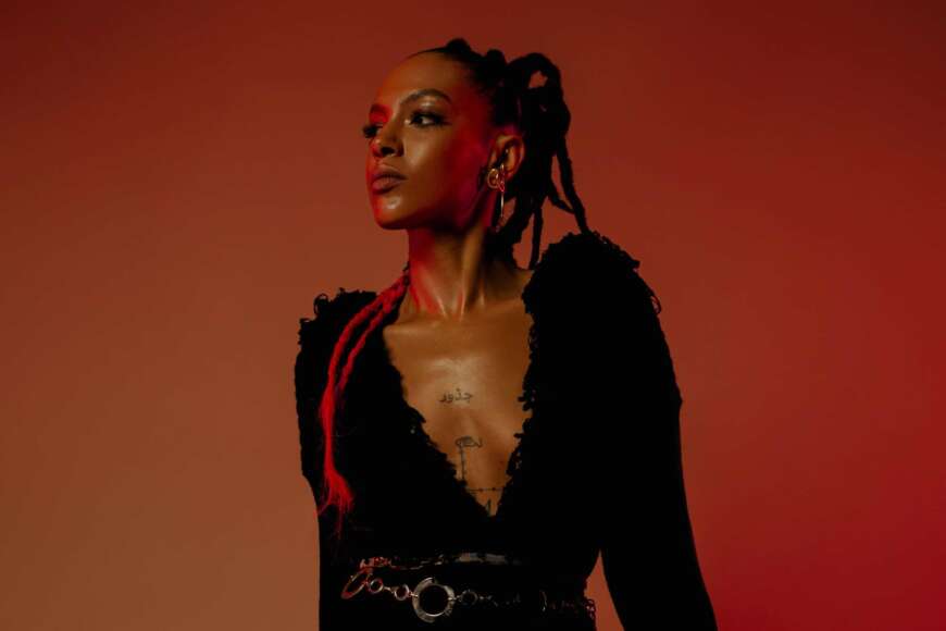 Alewya steht vor einem orangefarbenen Hintergrund. Sie trägt ein tief ausgeschnittenes, schwarzes Oberteil.