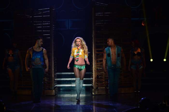 Britney Spears ein gelbes Oberteil und eine kurze grüne Hose. Links und rechts von ihr stehen zwei männliche Tänzer.