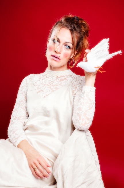 Charlotte Brandi sitzt in einem weißen Kleid vor einem roten Hintergrund und hält eine weiße Stofftaube in der linken Hand.