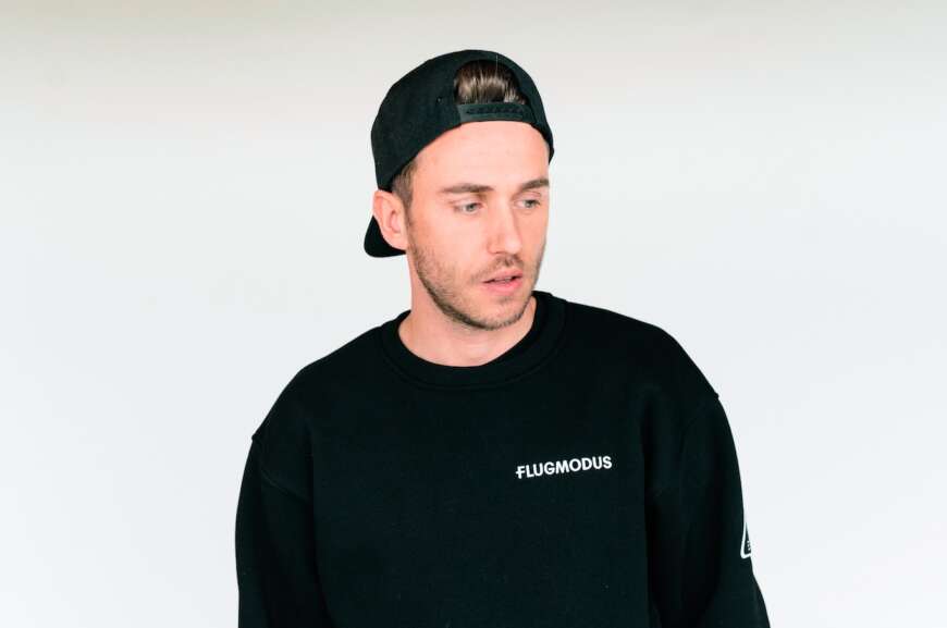 Clueso steht in einem schwarzen Pullover und einer Basecap vor einem hellen Hintergrund. Von der Brust aufwärts fotografiert.