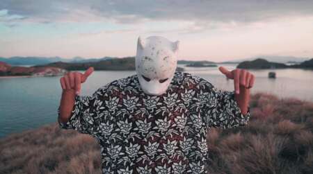 Cro steht mit Maske und in einem T-Shirt auf einer Insel im Meer und schaut nach unten. Mit seinen Zeigefingern zeigt er ebenfalls nach unten.
