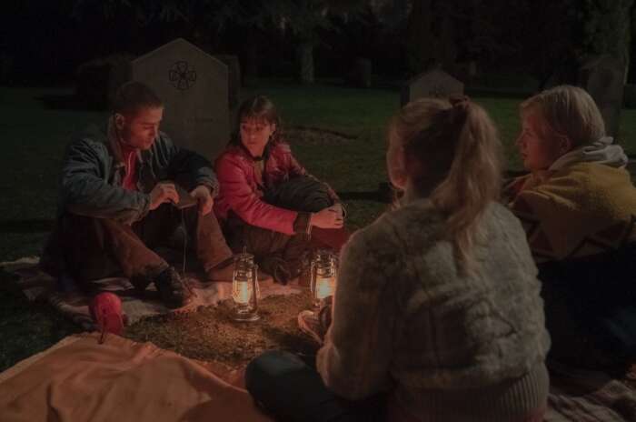 Bild aus der Serie „Equinox“: Vier Jugendliche sitzen sich mit Laternen auf dem Boden gegenüber. Die Umgebung lässt auf einen Friedhof schließen.