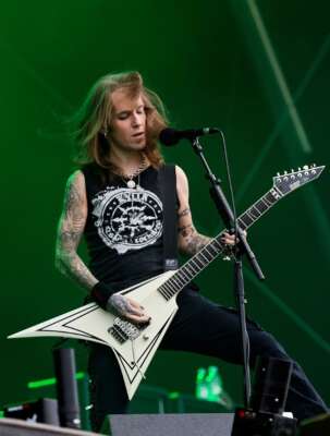 Alexi Laiho steht mit seiner Gitarre vor einem grünen Hintergrund.