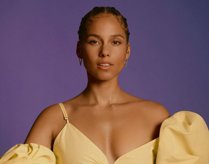Alicia Keys steht in einem gelben Oberteil vor einem violetten Hintergrund. Das Bild zeigt die Künstlerin von der Körpermitte aufwärts.