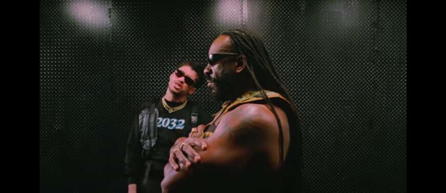 Bad Bunny steht mit Booker T in einem dunklen Raum. Beide Tragen Sonnenbrillen, Booker T starrt mit verschränkten Armen nach vorne, während Bad Bunny in die Kamera guckt.