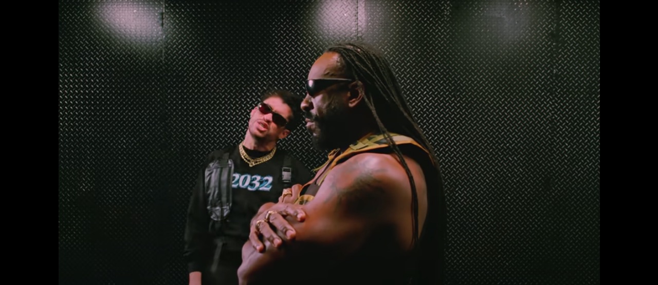 Bad Bunny steht mit Booker T in einem dunklen Raum. Beide Tragen Sonnenbrillen, Booker T starrt mit verschränkten Armen nach vorne, während Bad Bunny in die Kamera guckt.