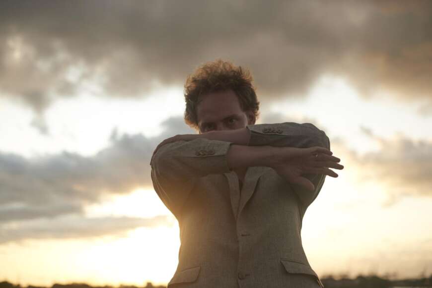 Karwendel steht mit verschränkten Armen vor dem Gesicht vor einer Wolkenfront. Im Hintergrund geht die Sonne unter.