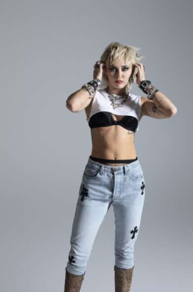 Miley Cyrus steht mit einer blauen Jeans, kurzen blonden Haaren und einem bauchfreien Top vor einem grauen Hintergrund.
