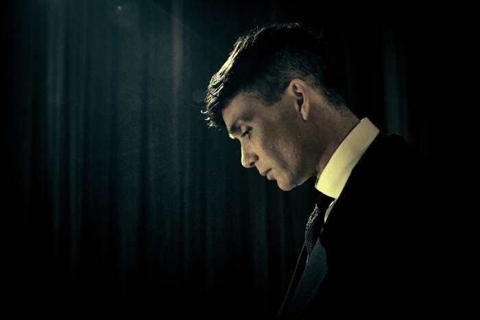 Ein Szenenbild aus „Peaky Blinders“: Thomas Shelby (Cillian Murphy) steht mit gebeugtem Kopf vor einem dunklen Hintergrund.