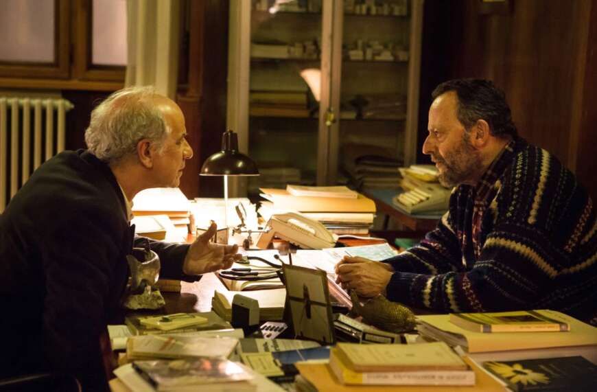 "Der Nebelmann": Vogel (Toni Servillo) und Flores (Jean Reno) sitzen sich an einem mit Büchern und Unterlagen überdeckten Schreibtisch gegenüber und sprechen miteinander.