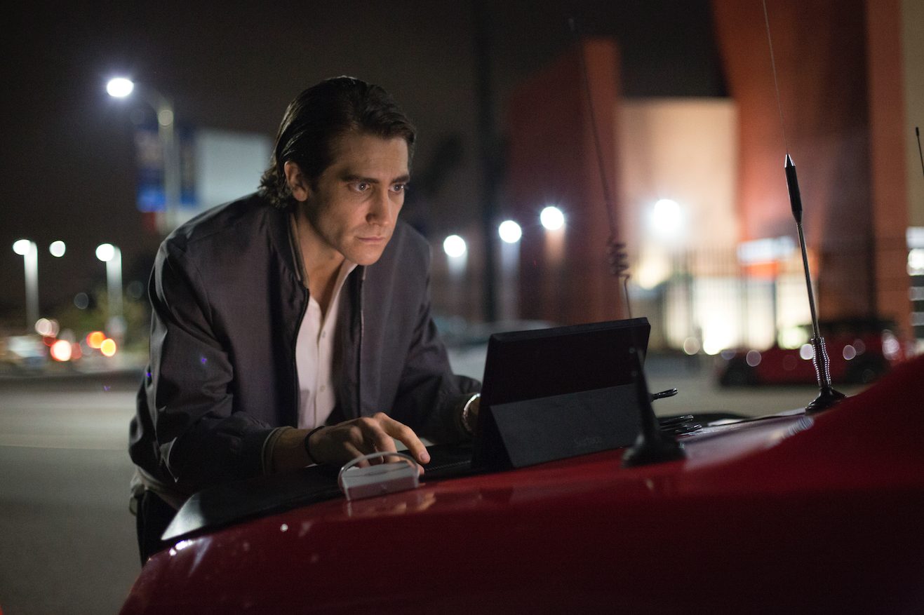 Szenenbild aus Nightcrawler: Lou Bloom (Jake Gyllenhaal) tippt auf einen Laptop auf der Kühlerhaube eines roten Autos, im Hintergrund das nächtliche LA.