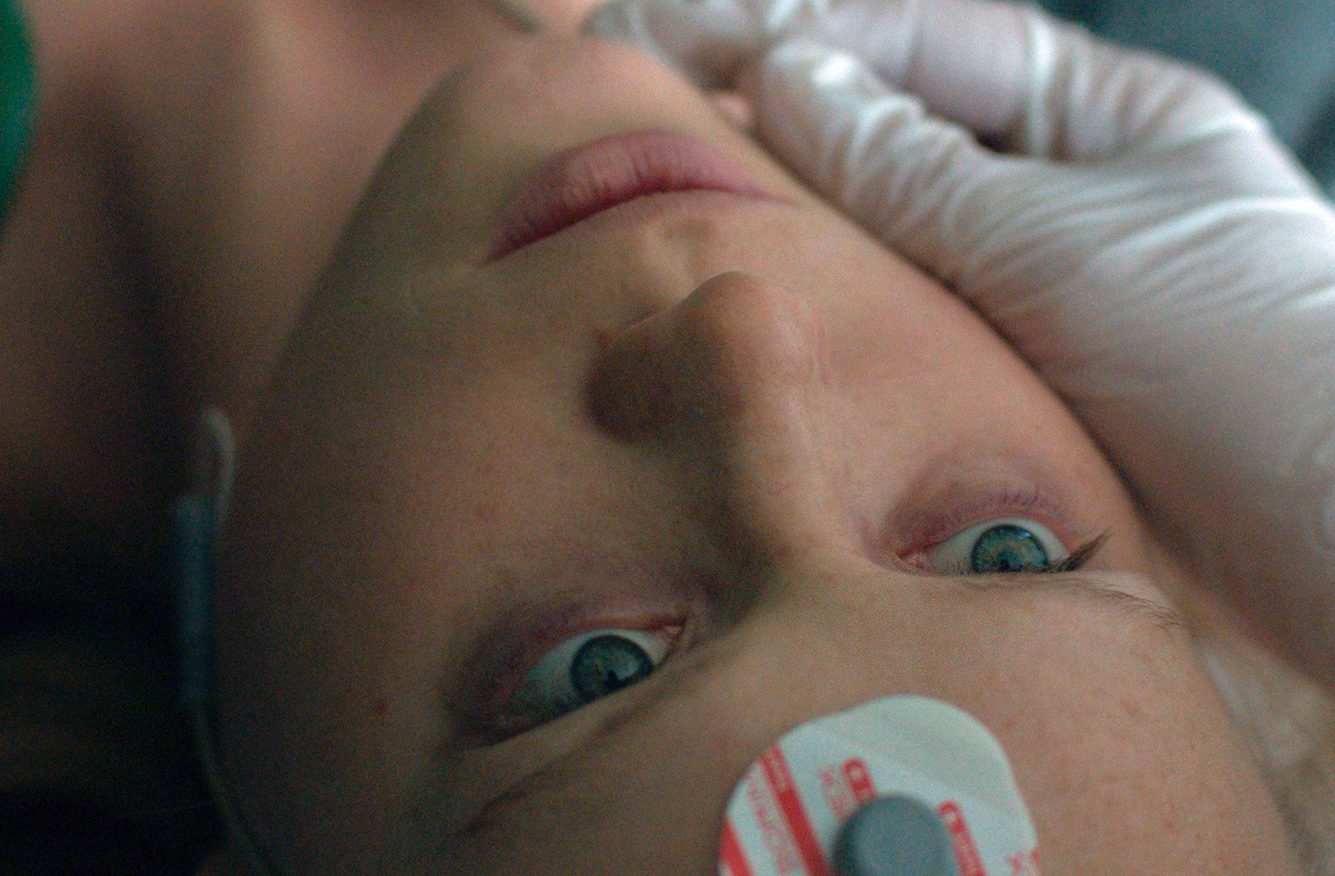 Szenenbild aus „Ohne ein Wort zu sagen“: Das Gesicht eines Mädchens mit einer Elektrode auf der Stirn wird von einer Hand mit weißen Handschuh fixiert.