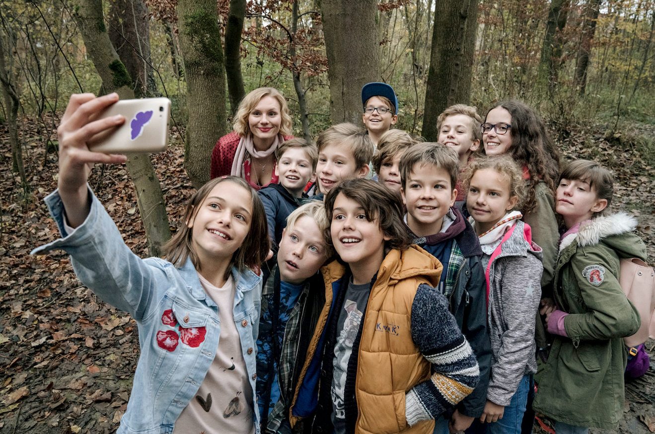 Szenenbild aus „Rufmord“: Eine Gruppe Schüler:innen und eine Lehrerin lächeln für einen Selfie in die Handykamera. Im Hintergrund der Wald.