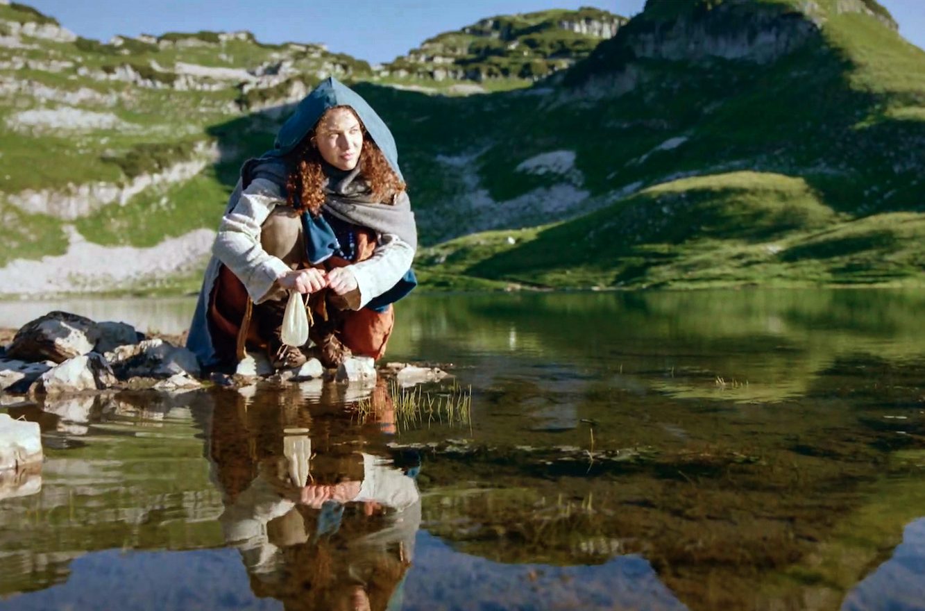 Die Salzfürstin Szenebild: Eine junge Frau in altertümlicher Kluft kniet an einem Bergsee mit einem weißen Beutel in der Hand.