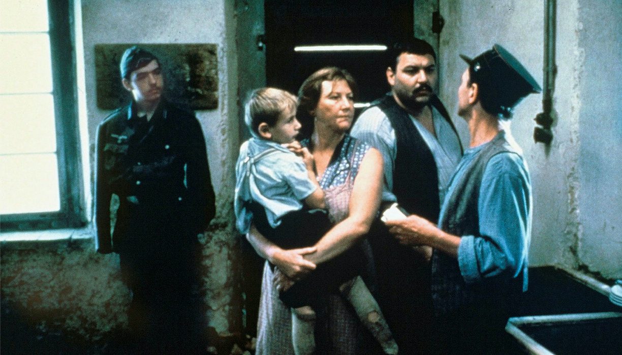 Ein Paar steht vor einem Mann mit Uniformsmütze, die Frau hat einen kleinen Jungen auf dem Arm. Im Hintergrund schaut ein Soldat zu.