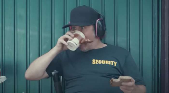Ein Mann mit einer Cap und Kopfhörern trinkt Kaffee aus einem Plastikbecher in seiner rechten Hand. In seiner linken hält er ein angebissenes Butterbrot. Er sitzt vor einem Garagentor. Auf seinem T-Shirt steht „Security“.
