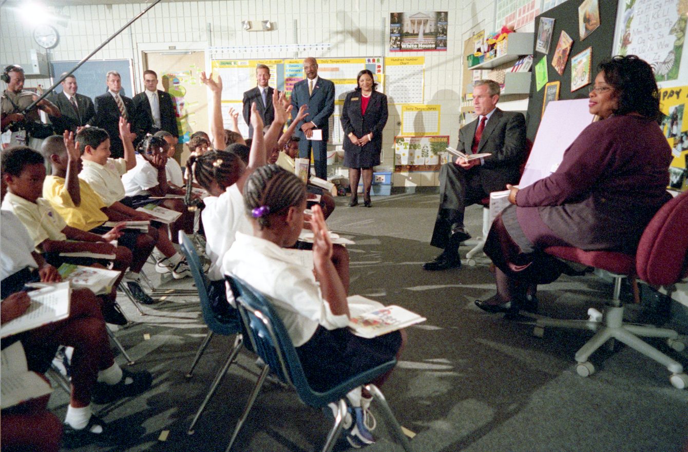 Ein Mann (George W. Bush) sitzt mit einem Buch auf dem Schoß in einem Klassenzimmer. Vor ihm im Halbkreis eine Gruppe Kinder in weißen Hemden. Ein Boom Mic hängt über ihren Köpfen.