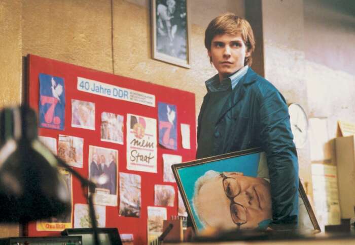 Ein junger Mann steht vor einer Pinnwand, auf der unter anderem zu lesen ist: „40 Jahre DDR“ und „Mein Staat“. Unter dem Arm trägt er ein Bild von Erich Honecker.