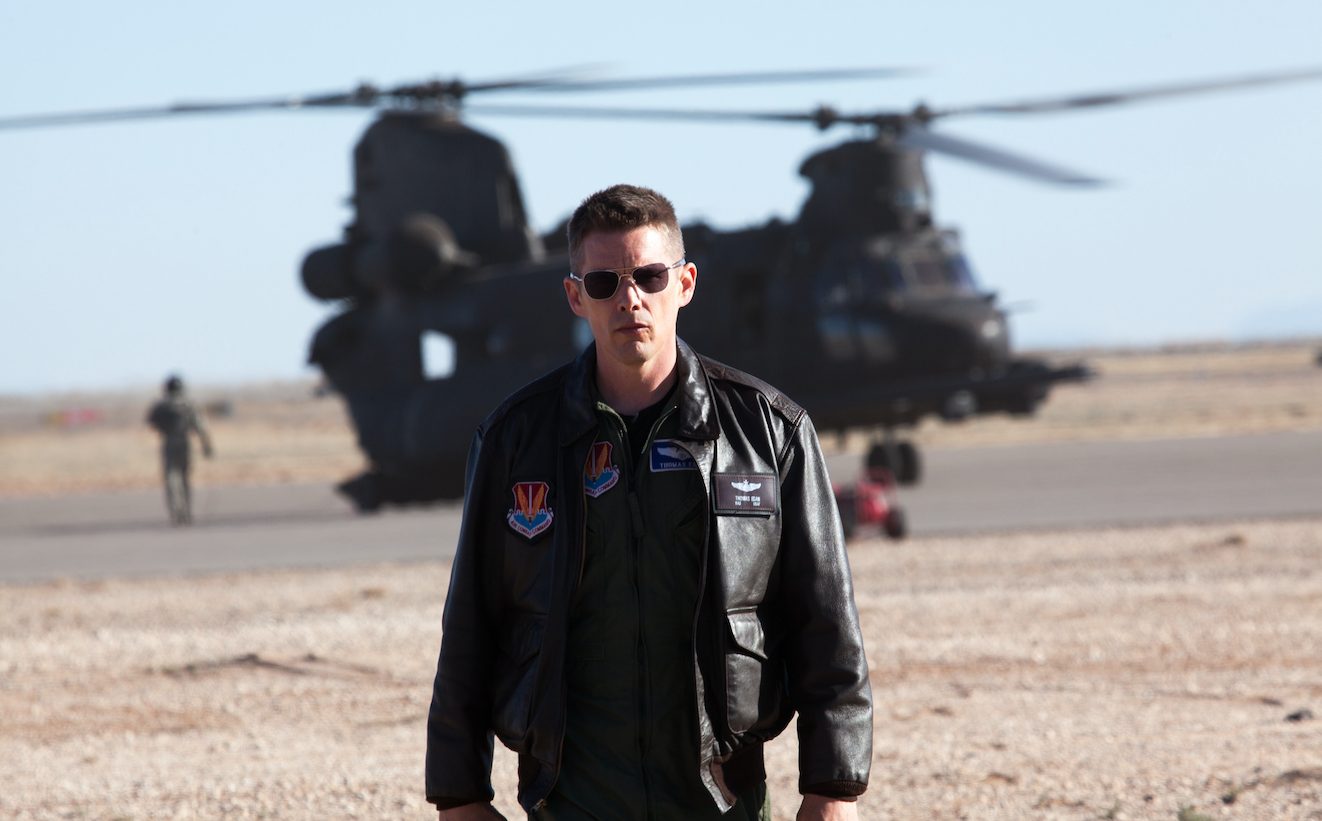 Ein Soldat in schwarzer Lederjacke und Sonnenbrille steht in einer Wüste, im Hintergrund ist ein Militär-Helikopter zu sehen.