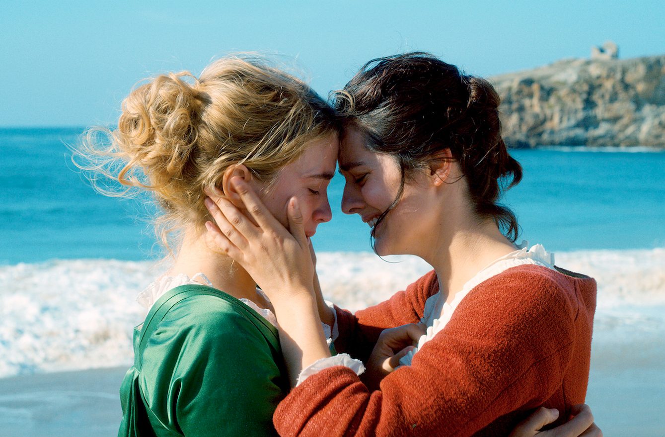 Zwei junge Frauen an einem Strand. Die rechte, in rotem Kleid mit dunklen Haaren, hält das Gesicht der rechten, im grünen Kleid mit blonden Haaren, in den Händen. Beide weinen.