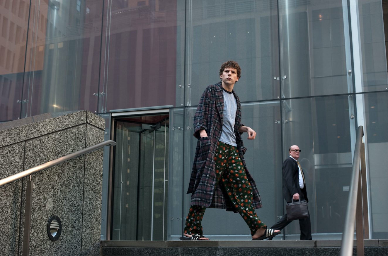 Ein junger Mann, Jesse Eisenberg, steht auf einem Treppenabsatz vor einem Gebäude mit Glasfront. Er trägt Schlafanzug, Bademantel und Flip-Flops.