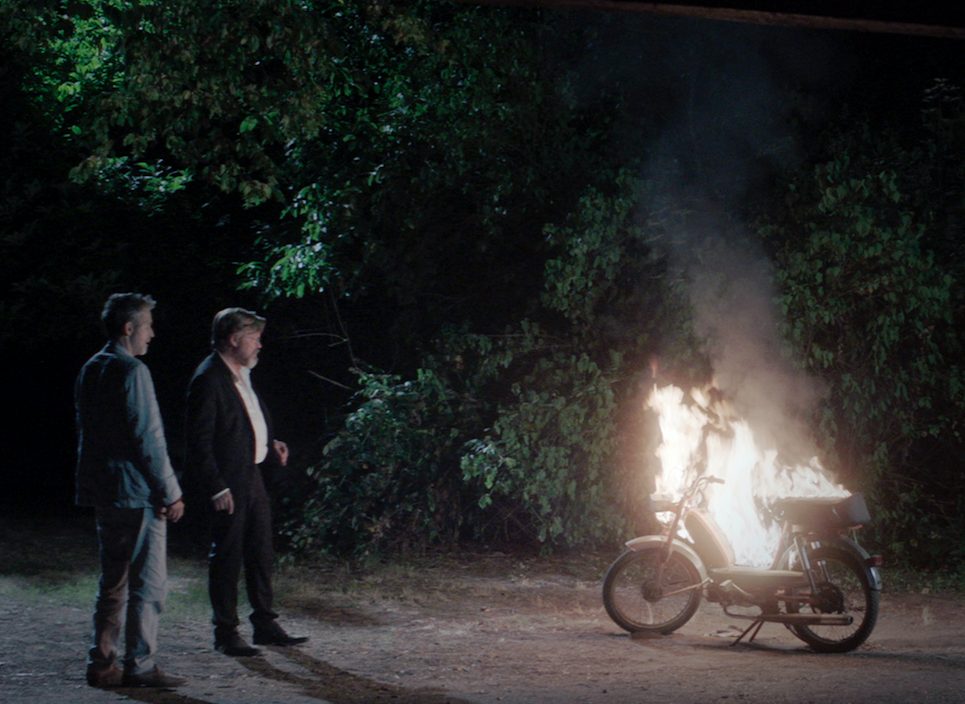 Zwei Männer in Anzügen stehen vor einem brennenden Mofa. Es ist Nacht, hinter ihnen Bäume.