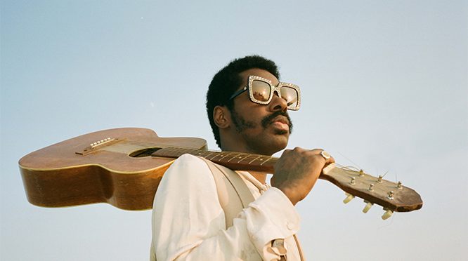 Curtis Harding hat eine glamouröse Sonnenbrille auf und seine Gitarre lässig über der Schulter.