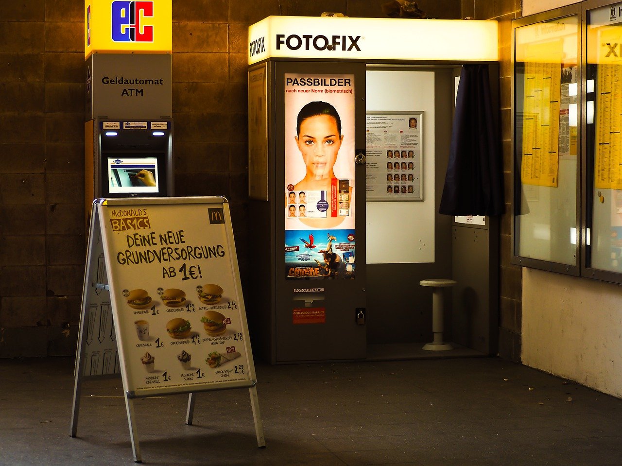 Ein typischer Fotoautomat, der neben einem Geldautomaten aufgestellt wurde. Davor ein Aufsteller mit Werbung.