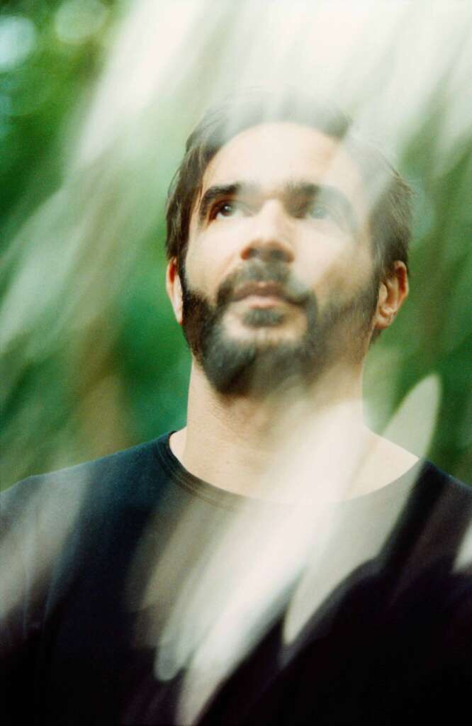 Jon Hopkins zwischen verschwommen, grünen Blättern in einem schwarzen T-Shirt.