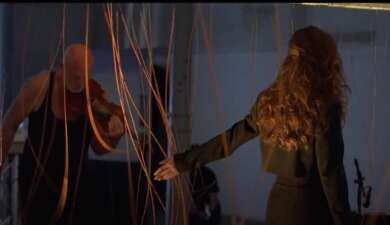 Abstrakte Linien und ein Geiger neben eine Schauspielerin mit langem Haar in einem Videostill aus einer Perfomance.