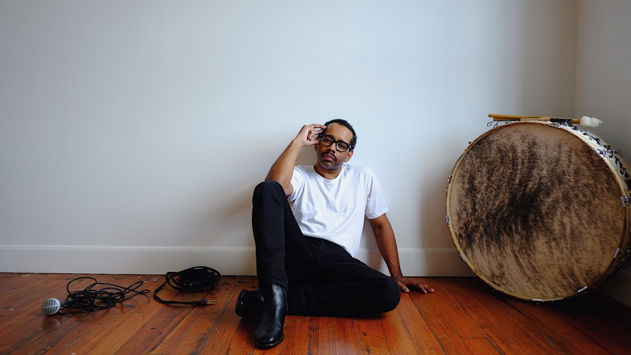 Jamire Williams sitzt auf einem Dielenboden vor einer weißen Wand neben einer großen, runden Trommel, einem Kabel und einem Mikro, die auf dem Boden liegen.