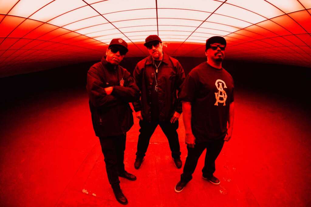Die drei Rapper von Cypress Hill posen in rotes Licht getaucht unter einem Fester und sind ganz in Schwarz gekleidet.