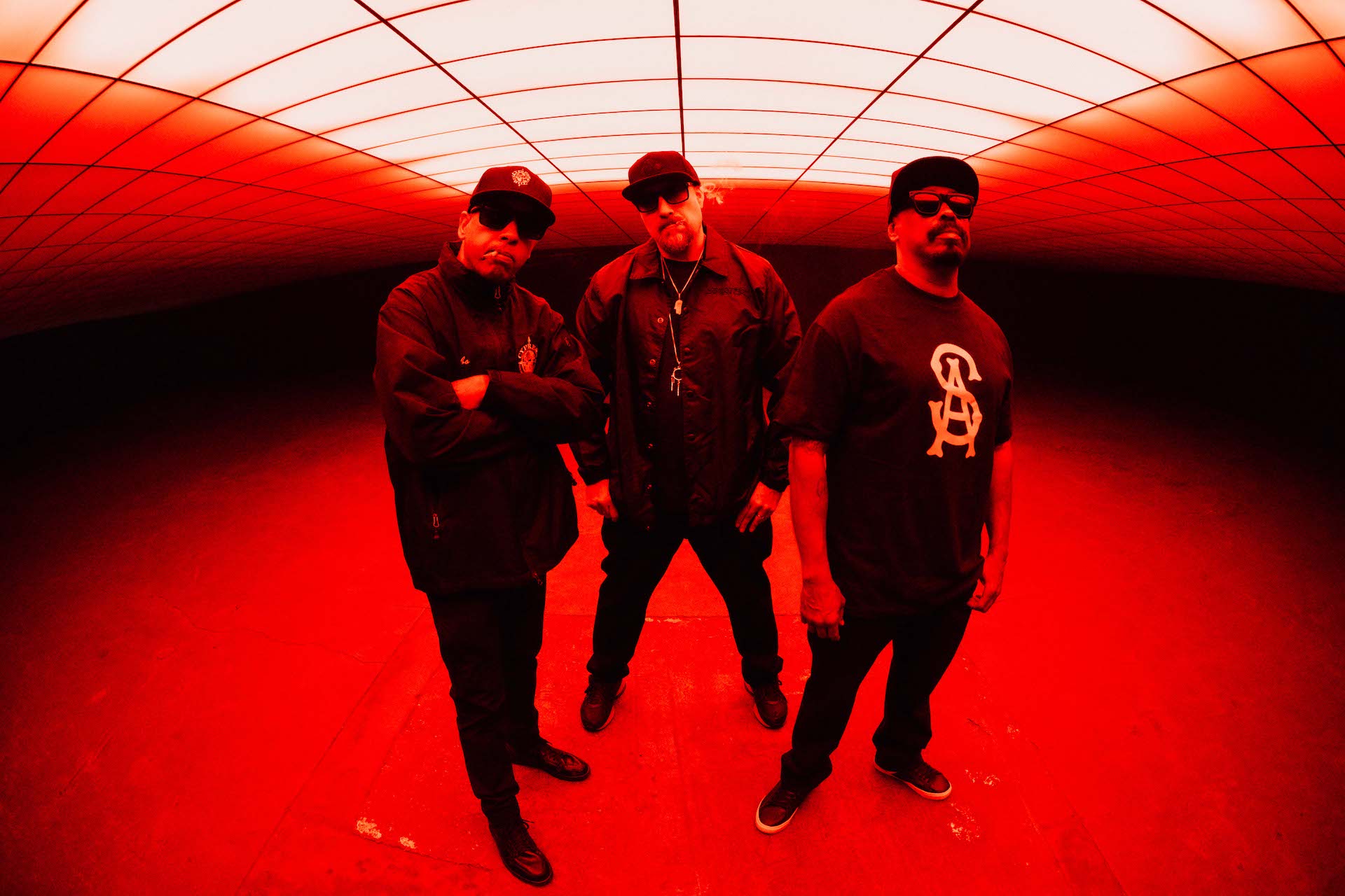 Die drei Rapper von Cypress Hill posen in rotes Licht getaucht unter einem Fester und sind ganz in Schwarz gekleidet.