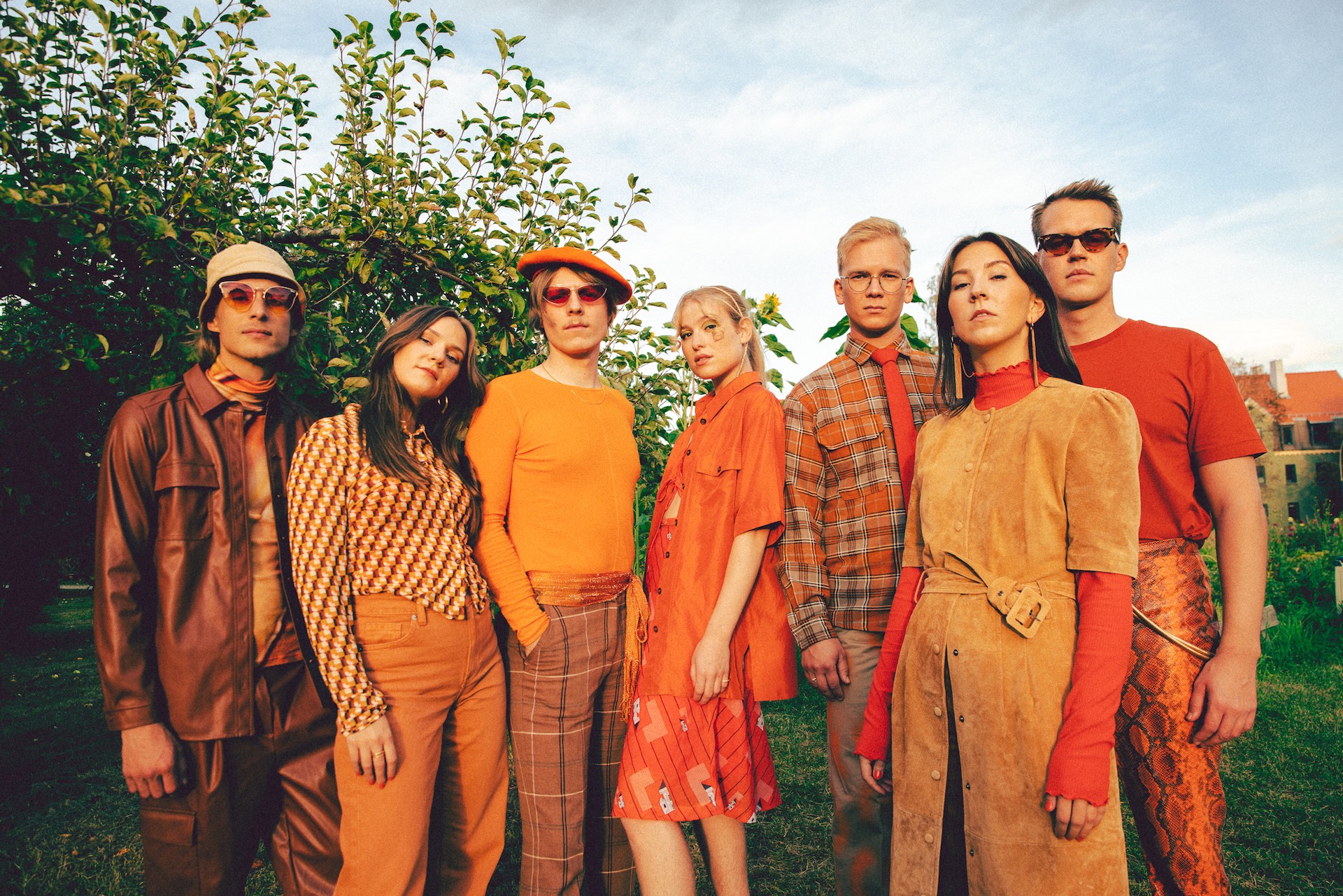 Alle sieben Mitgleider der Gruppe fieh stehen in orangefarbenen Outfits in einem stattgrünen Garten.