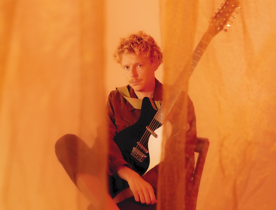 Peter the human boy sitzt mit seiner Gitarre auf einem Stuhl hinter einem leicht durchsichtigen Vorhang. Die Szenerie ist in orangenes Licht getaucht.