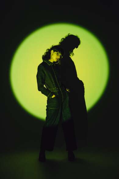 Die Musikerin C'est Karma trägt einen schwarzen Overall und posiert in einem grünen Spotlight.
