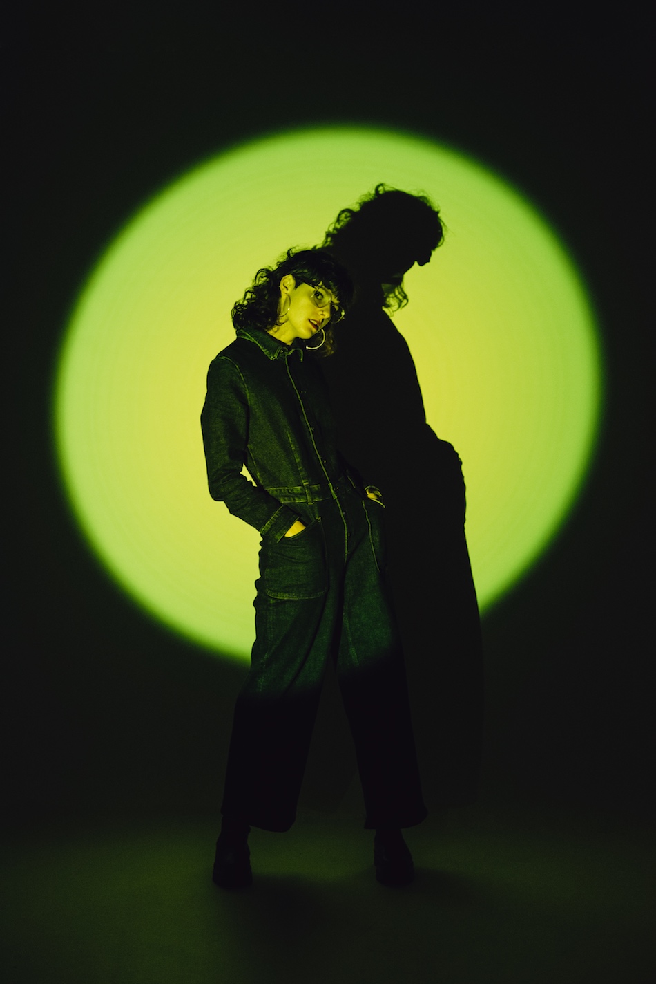 Die Musikerin C'est Karma trägt einen schwarzen Overall und posiert in einem grünen Spotlight.