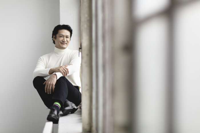 Jae-Hyuck Cho sitzt in weißem Pulli, schwarzen Hose und Schuhen, aber mit bunten Socken auf einer Fensterbank.