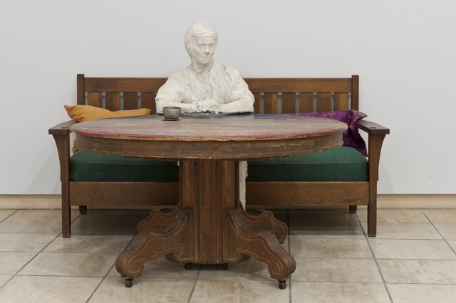 Ein weiße Statue namens Ruth sitzt auf einer alten Holzbank mit dunkelgrünem Sitzkissen an einem massiven Holztisch.