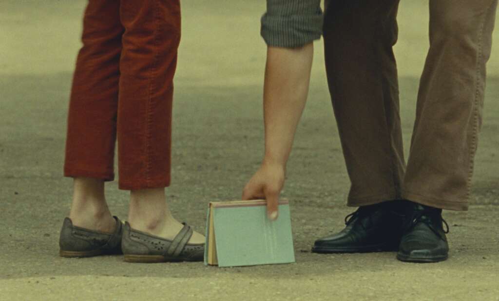 Zwei Paar Füße und eine Hand, die ein dazwischenliegendes Buch hochhebt sind zu sehen. - Filmstill aus „Was sehen wir, wenn wir zum Himmel schauen?“