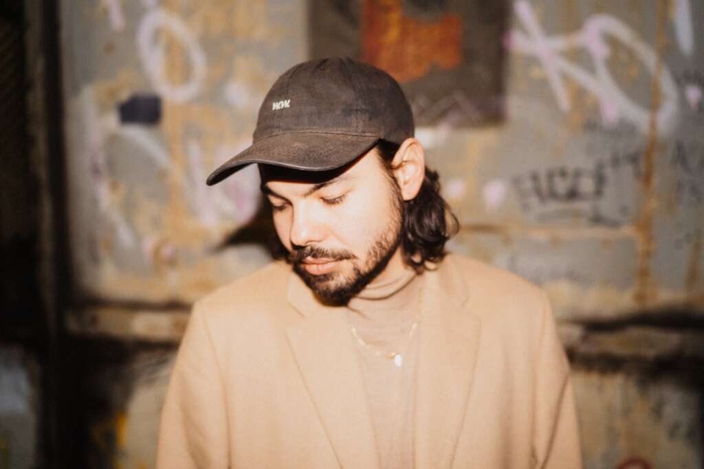Der Musiker Anoki ist in einem beigen Rollkragenpullover und mit schwarzer Cap vor einer mit Graffiti-Tag bemalten Wans zu sehen.