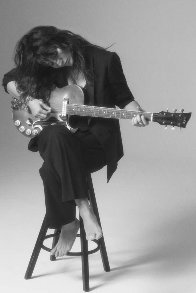 Zu sehen ist eine Schwarz-weiß-Aufnahme von Chiara Civello im Schwarzen Anzug, auf einem barhocker sitzend, auf einer Gitarre spielend.