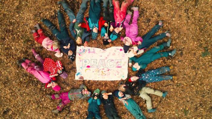 Eine Gruppe lachender Kinder ist zu sehen, die im Kreis auf buntem Laub um ein Banner mit der Aufschrift „Peace“ liegt.
