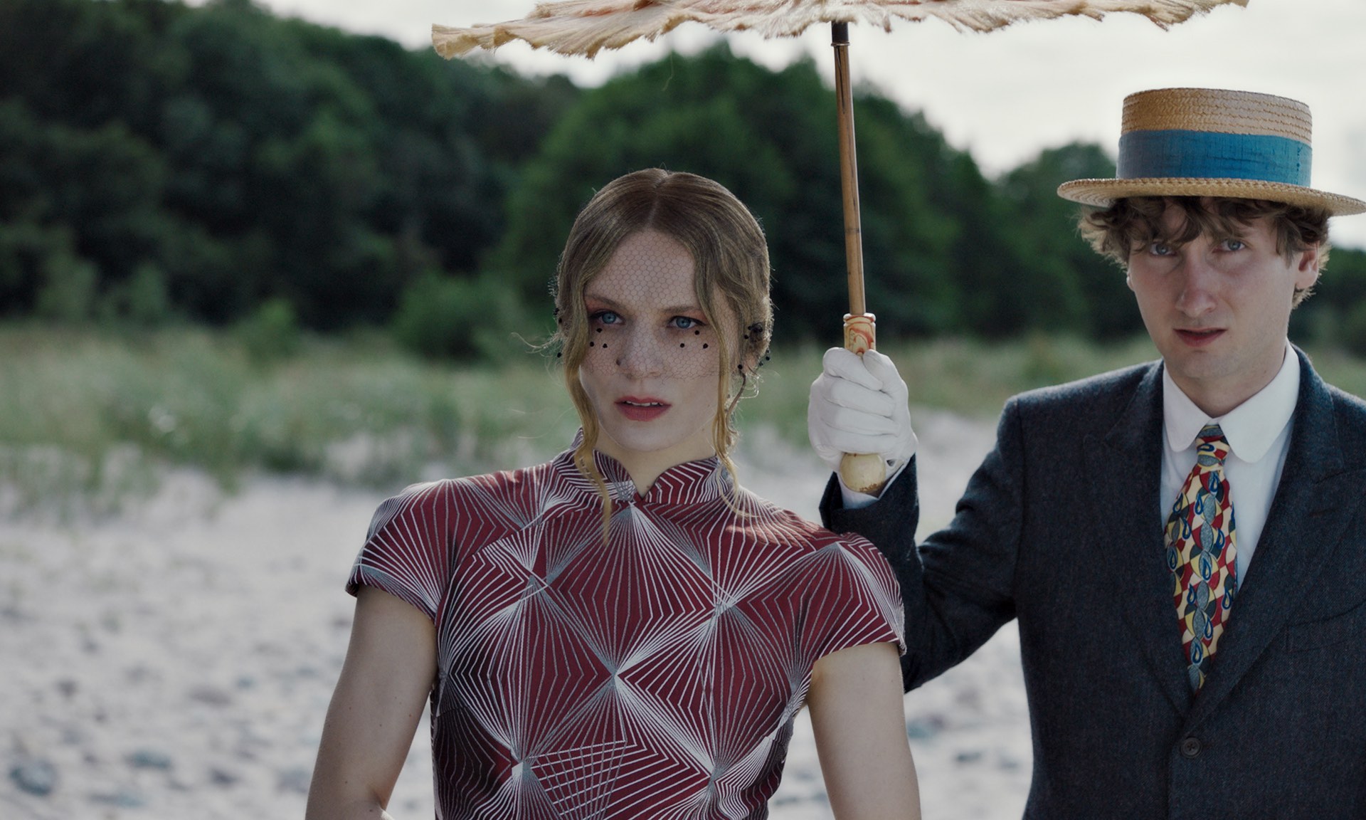 Zu sehen ist ein Still aus dem Film „Blutsauger“ auf dem zwei blasse, vampirmäßig aussehende Personen am Strand erschrocken in die kamera schauen, während sie sich mit einem Schirm in der Hand vor der Sonne schützen.