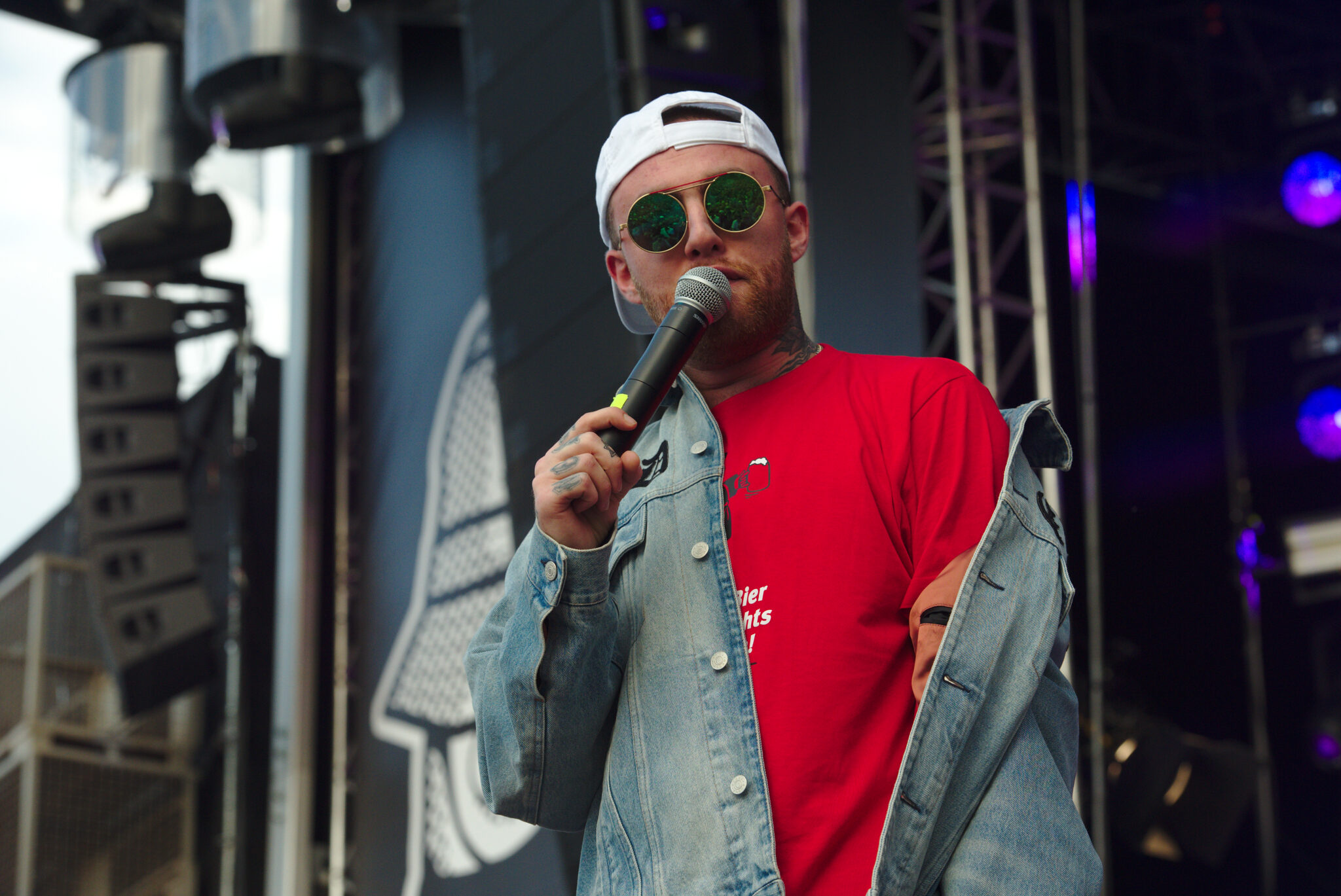 Rapper Mac Miller auf der Bühne mit roten T-Shirt, Jeansjacke, weißem Cappie, Sonnenbrille und Mikro vor dem Mund.